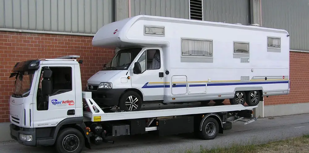 Camping-car capucine chargé sur le plateau d'un camion de dépannage, devant un hangar