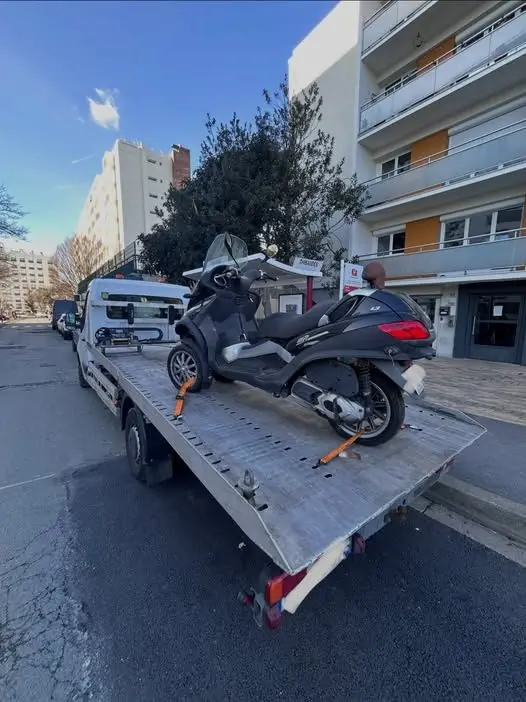 Scooter trois-roues sanglé sur le plateau abaissé d'un camion de dépannage, en pied d'immeuble